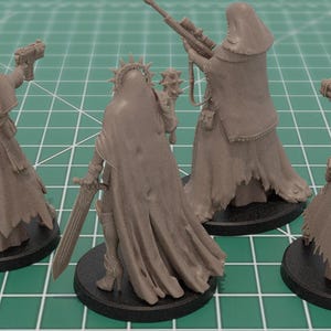 Op de afbeelding: Vier ongeverfde grijze miniatuurfiguren van hars op ronde zwarte voetstukken. Elke figuur draagt een kapmantel en draagt een wapen, waaronder een pistool, een strijdhamer, een sluipschuttersgeweer en een kanon, met gedetailleerde stoftexturen.