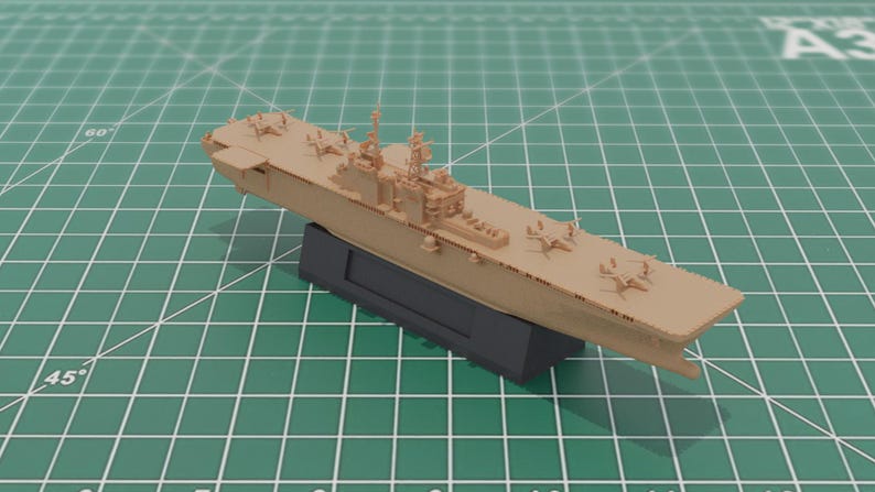 May include: A tan model aircraft carrier with miniature aeroplanes on the deck, mounted on a black base. The model is set against a green grid background. The model is a detailed replica.