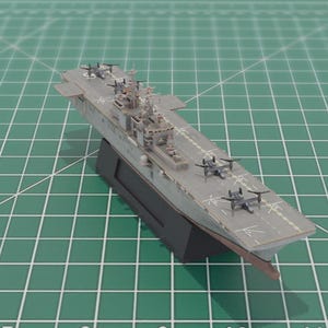 May include: A scale model of a grey aircraft carrier, complete with miniature aeroplanes. The model is mounted on a black stand, set against a green cutting mat. The carrier features detailed elements such as the control tower and deck markings.
