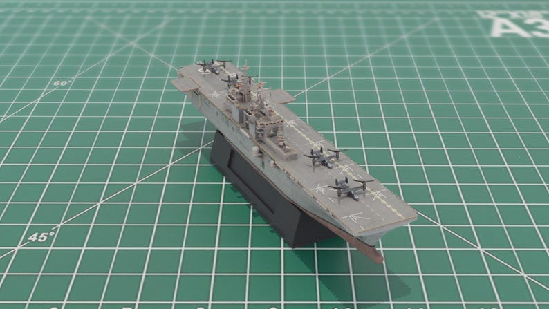 May include: A scale model of a grey aircraft carrier, complete with miniature aeroplanes. The model is mounted on a black stand, set against a green cutting mat. The carrier features detailed elements such as the control tower and deck markings.