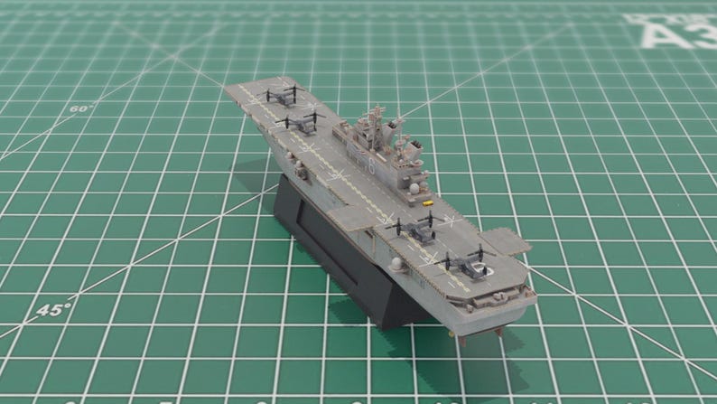 May include: A detailed model of a grey aircraft carrier with a black base. The model features miniature helicopters and other aircraft on the flight deck. The carrier is set against a green grid background.
