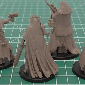 Op de afbeelding: Vier onbeschilderde grijze miniatuurfiguren van hars op zwarte ronde voetstukken. Elk figuur draagt een mantel met capuchon en is uitgerust met verschillende voorwerpen, waaronder een pistool, een zwaard, een strijdhamer, een sluipschuttersgeweer en een raketwerper.
