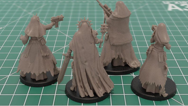 Op de afbeelding: Vier onbeschilderde grijze miniatuurfiguren van hars op zwarte ronde voetstukken. Elk figuur draagt een mantel met capuchon en is uitgerust met verschillende voorwerpen, waaronder een pistool, een zwaard, een strijdhamer, een sluipschuttersgeweer en een raketwerper.