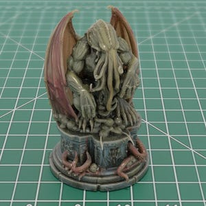 May include: A hand-painted miniature figurine of a Cthulhu-like creature with tentacles, wings, and a muscular build, perched on a circular stone-textured pedestal base with carved details and small tentacle accents in shades of green and brown.