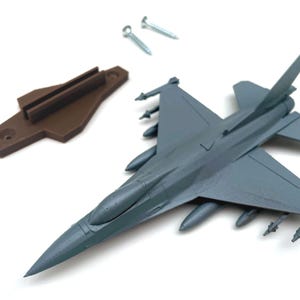 May include: A gray plastic model fighter jet with mounted missiles, accompanied by a brown plastic mounting bracket and two small silver metal screws. The aircraft features a pointed nose, swept wings, and a vertical stabilizer on the tail section.