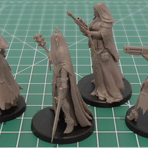 Op de afbeelding: Vier ongeverfde grijze hars miniatuurfiguren op ronde zwarte voetstukken. Elk figuur draagt een kapmantel en tactische uitrusting. De figuren zijn uitgerust met diverse wapens, waaronder een pistool, een strijdhamer, een geweer en een zwaar vuurwapen.