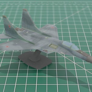 May include: A gray model jet fighter with red star markings and the number "385" on the side. The model is mounted on a black stand and sits on a green grid surface.