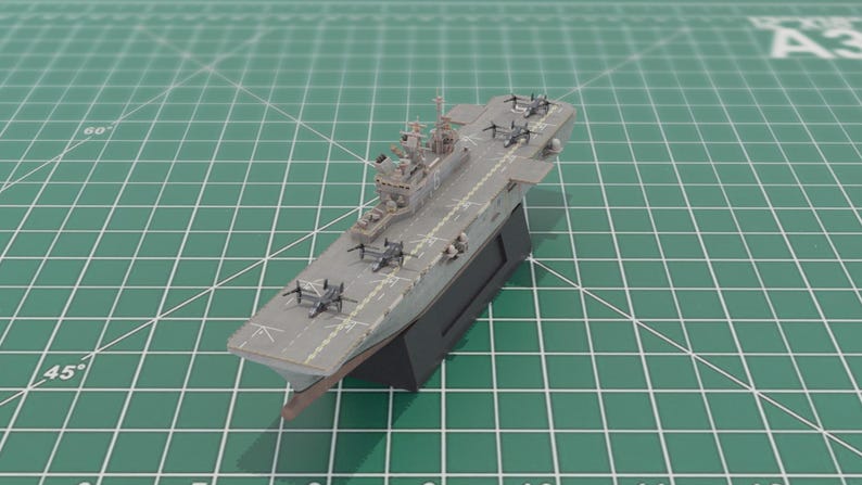 May include: A detailed model of a grey aircraft carrier with miniature aeroplanes on the deck. The model is mounted on a black base against a green grid background. The carrier features intricate details.