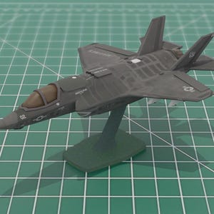 May include: A dark gray model airplane, possibly an F-35 fighter jet, mounted on a green stand. The model has detailed features, including a cockpit and wings. The background is a green grid.