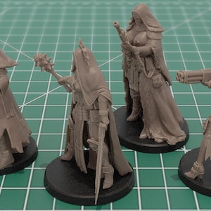 Op de afbeelding: Vier onbeschilderde miniatuurfiguren van grijze hars op ronde zwarte voetstukken. Elk figuur draagt een kapmantel en tactische uitrusting. De figuren zijn uitgerust met diverse wapens, waaronder een pistool, een strijdhamer, een geweer en een kanon.