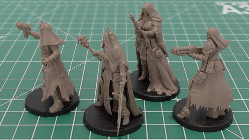 Op de afbeelding: Vier onbeschilderde miniatuurfiguren van grijze hars op ronde zwarte voetstukken. Elk figuur draagt een kapmantel en tactische uitrusting. De figuren zijn uitgerust met diverse wapens, waaronder een pistool, een strijdhamer, een geweer en een kanon.