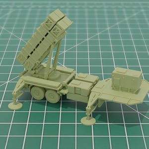 May include: A light green model of a military missile launcher on a wheeled platform. The launcher has a rectangular structure with multiple compartments and is set against a green grid background.