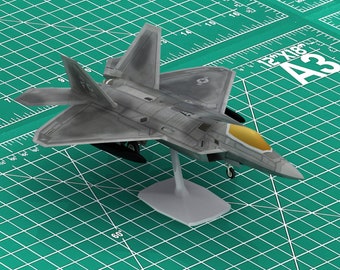 May include: A grey and gold model aircraft on a white stand. The model is of an F-22 Raptor fighter jet. The model is on a green cutting mat with a ruler and the text "12"x18" A3" printed on it.