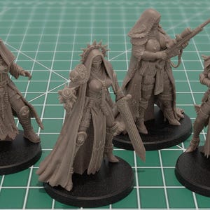 Op de afbeelding: Vier onbeschilderde miniatuurfiguren van grijze hars op ronde zwarte voetstukken. Elk figuur draagt een harnas met capuchon en draagt een ander wapen, waaronder een pistool, een zwaard, een sluipschuttersgeweer en een zwaar wapen met meerdere lopen.
