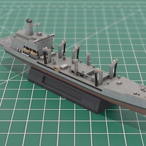 Może przedstawiać: Szary model statku o szczegółowym projekcie, z nadbudówką, żurawiami i kadłubem. Model jest zamontowany na czarnej podstawie i spoczywa na zielonej powierzchni kratki. Długość statku to około 25 cm.