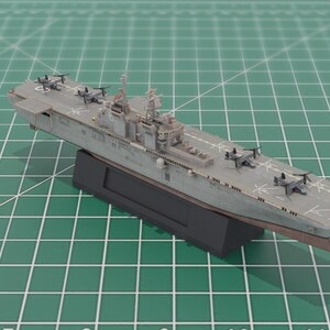 May include: A grey model aircraft carrier with miniature aircraft on the deck. The model is mounted on a black base and is positioned on a green grid surface. The model is approximately 30 cm long.