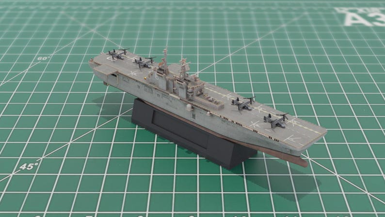 May include: A grey model aircraft carrier with miniature aircraft on the deck. The model is mounted on a black base and is positioned on a green grid surface. The model is approximately 30 cm long.