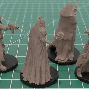 Op de afbeelding: Vier onbeschilderde grijze hars miniatuurfiguren op ronde zwarte voetstukken. De figuren dragen lange gewaden, sluiers en tactische uitrusting, waarbij er drie futuristische vuurwapens vasthouden en &eacute;&eacute;n een strijdknots met stekels in de hand houdt.