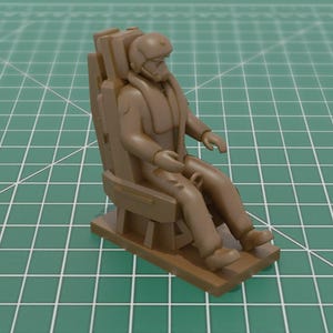 Peut inclure: Figurine miniature marron d'un pilote assis dans un siège éjectable. Le pilote porte un casque et une combinaison de vol. Le siège a une base rectangulaire et un dossier détaillé. La figurine est posée sur un fond quadrillé vert.