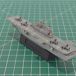May include: A grey model aircraft carrier with miniature aircraft on the deck. The model is mounted on a black base, resting on a green cutting mat. The carrier has a detailed design.