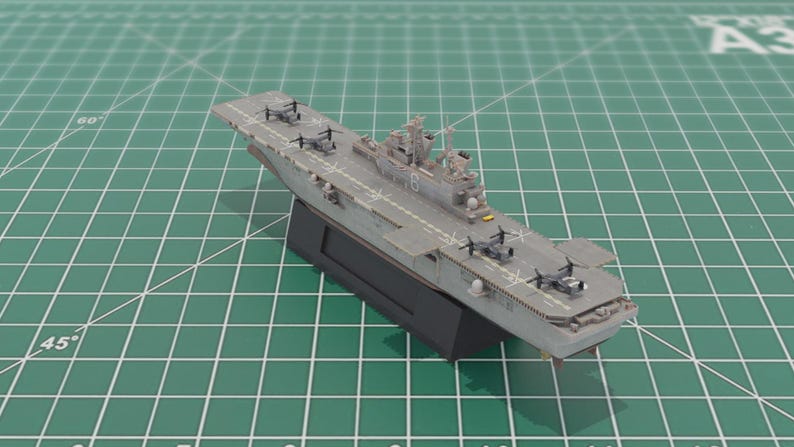 May include: A grey model aircraft carrier with miniature aircraft on the deck. The model is mounted on a black base, resting on a green cutting mat. The carrier has a detailed design.