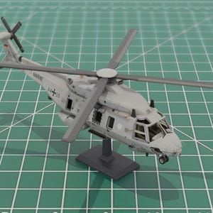 May include: A detailed model of a military helicopter, painted in shades of gray, with a four-blade main rotor and a tail rotor. The model is mounted on a small stand, set against a green grid background. The word "MARINE" is visible on the side.