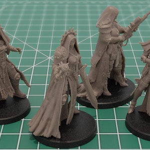 Op de afbeelding: Vier ongeverfde lichtbruine hars miniatuurfiguren op ronde zwarte voetstukken. Elk figuur stelt een krijger voor in een mantel met capuchon en harnas, met diverse wapens waaronder een geweer, een zwaard en een zwaar vuurwapen in actie.