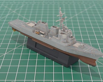 May include: A gray model warship with a brown hull, mounted on a black base. The ship features detailed structures, including a radar mast and a helipad. The model is set against a green grid background.