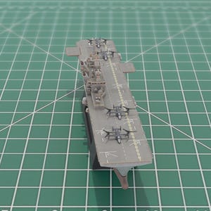 May include: A grey model aircraft carrier with miniature aeroplanes on the deck. The model is positioned on a green cutting mat with grid lines, highlighting the carrier's design and features.