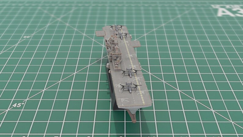 May include: A grey model aircraft carrier with miniature aeroplanes on the deck. The model is positioned on a green cutting mat with grid lines, highlighting the carrier's design and features.
