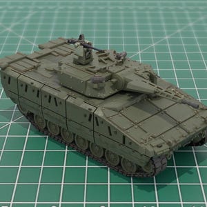 May include: An olive green model tank with a long gun barrel and multiple turrets. The tank sits on a green grid surface, showcasing its detailed design. The tank's tracks and overall structure are clearly visible.