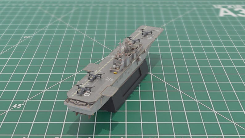 May include: A grey model aircraft carrier with miniature aircraft on the deck, set on a black base. The model is positioned on a green grid surface. The number 6 is visible on the deck of the carrier.