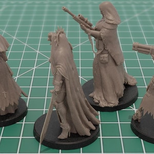 Op de afbeelding: Vier ongeverfde grijze hars miniatuurfiguren op ronde zwarte voetstukken. De figuren dragen lange capuchonmantels en dragen diverse uitrusting, waaronder een geweer met lange loop, een zwaard en een zwaar vuurwapen, geplaatst op een groene rastermat.