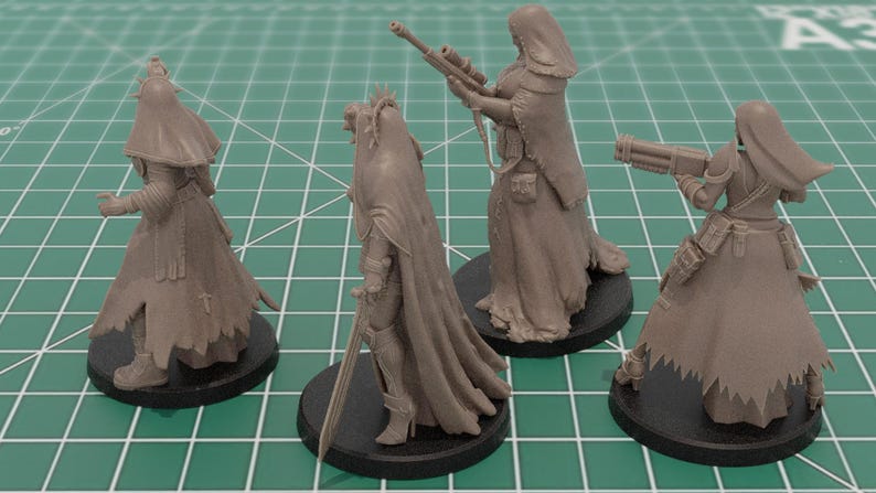 Op de afbeelding: Vier ongeverfde grijze hars miniatuurfiguren op ronde zwarte voetstukken. De figuren dragen lange capuchonmantels en dragen diverse uitrusting, waaronder een geweer met lange loop, een zwaard en een zwaar vuurwapen, geplaatst op een groene rastermat.