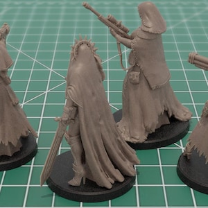 Op de afbeelding: Vier ongeverfde grijze hars miniatuurfiguren op ronde zwarte voetstukken. Elk figuur draagt een mantel met capuchon en een lange rok. De figuren zijn uitgerust met diverse wapens, waaronder een zwaard, een geweer en een zwaar kanon.