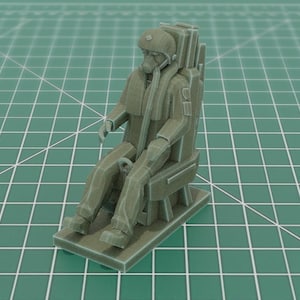 May include: A gray 3D printed miniature figurine of a pilot seated in an ejection seat. The pilot wears a flight suit, helmet, and oxygen mask with a hose connected to the chest. The figure is mounted on a rectangular base with visible layer lines.