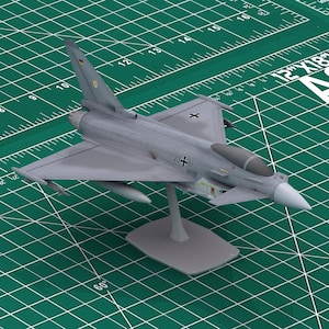 May include: A detailed model of a gray military jet fighter, mounted on a gray stand. The model has a white nose cone and is set against a green grid background. The model has the number "21+51" and a German flag on the tail.