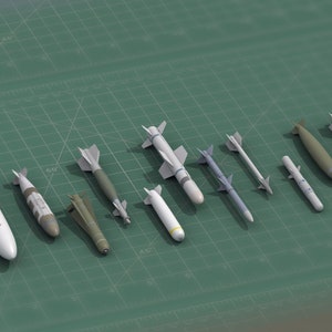 May include: A collection of nine miniature scale model missiles and bombs arranged in a row. The set includes various shapes and sizes, featuring white, grey, and olive green finishes, with some models displaying small fins and distinct nose cones.