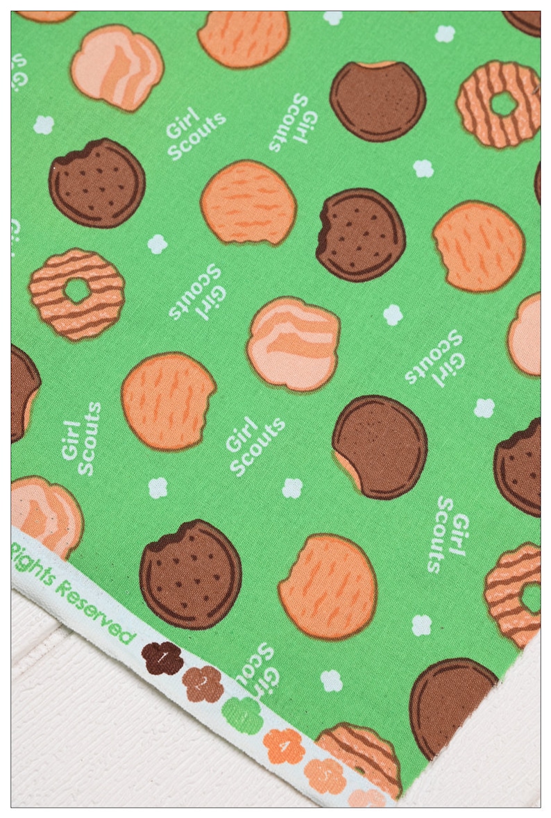 Może przedstawiać: Zielony materiał z powtarzającym się wzorem ciasteczek Girl Scout. Ciasteczka są w r&oacute;żnych kształtach i kolorach, w tym czekoladowe, kruche i z masłem orzechowym. Tekst "Girl Scouts" jest powtarzany na całym materiale.