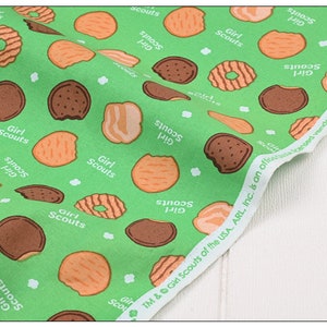 Może przedstawiać: Zielony materiał z powtarzającym się wzorem ciasteczek Girl Scout. Ciasteczka są w r&oacute;żnych kształtach i kolorach, w tym czekoladowe, kruche i z masłem orzechowym. Materiał zawiera r&oacute;wnież tekst "Girl Scouts" i "S'mores 140".
