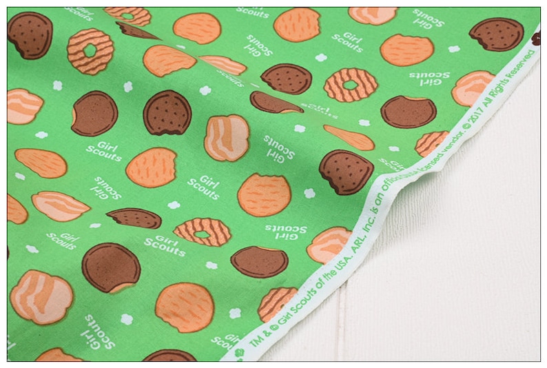 Może przedstawiać: Zielony materiał z powtarzającym się wzorem ciasteczek Girl Scout. Ciasteczka są w r&oacute;żnych kształtach i kolorach, w tym czekoladowe, kruche i z masłem orzechowym. Materiał zawiera r&oacute;wnież tekst "Girl Scouts" i "S'mores 140".