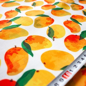 May include: White fabric with a repeating pattern of yellow and orange mangoes with green leaves. A white measuring tape with black markings is partially visible in the bottom right corner.