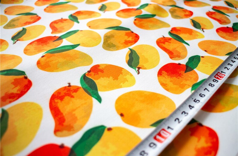 May include: White fabric with a repeating pattern of yellow and orange mangoes with green leaves. A white measuring tape with black markings is partially visible in the bottom right corner.