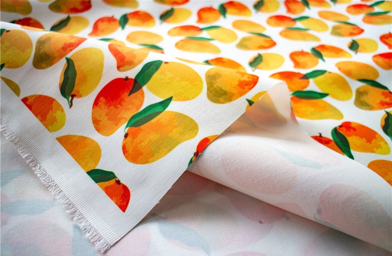 May include: White fabric with a repeating pattern of yellow and orange mangoes with green leaves. The fabric is folded over, showing the white underside.