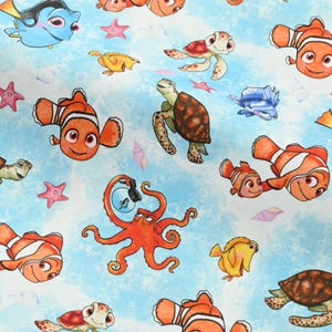 May include: A light blue fabric featuring cartoon illustrations of various sea creatures. The design includes orange clownfish, sea turtles, starfish, and an octopus. The background has a watercolor effect with bubbles and light blue hues.