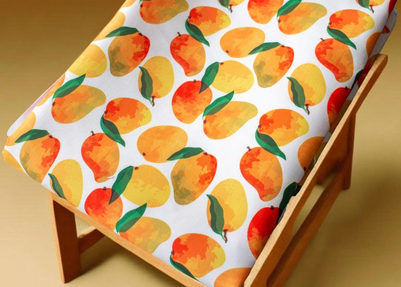 May include: A white fabric with a repeating pattern of yellow and orange mangoes. The fabric is draped over a wooden chair.