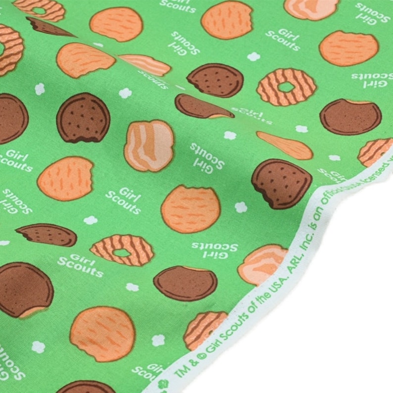 Może przedstawiać: Zielony materiał z powtarzającym się wzorem ciasteczek Girl Scout i tekstem "Girl Scouts". Ciasteczka są w r&oacute;żnych kształtach i kolorach, w tym czekoladowe, maślane i z masłem orzechowym.