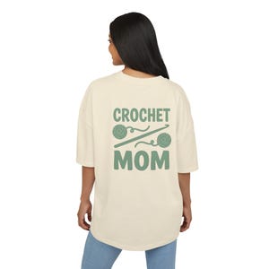 Könnte beinhalten: Ein übergroßes cremefarbenes T-Shirt mit einer salbeigrünen Grafik auf der Rückseite. Das Design enthält den Text "CROCHET MOM" in fetter Schrift, getrennt durch eine grafische Darstellung einer Häkelnadel zwischen zwei Wollknäueln.