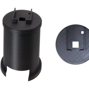 Könnte beinhalten: Ein schwarzes 3D-gedrucktes Objekt mit einer runden Basis und einem zylindrischen Oberteil. Das Oberteil hat eine quadratische Öffnung mit einer kleineren quadratischen Öffnung im Inneren. Das Objekt ist so konzipiert, dass es als Halter für einen kleinen Gegenstand verwendet werden kann.