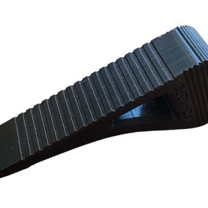 May include: A black rubber doorstop with a textured surface and a small hole in the center. The doorstop is shaped like a wedge and has a rounded top.